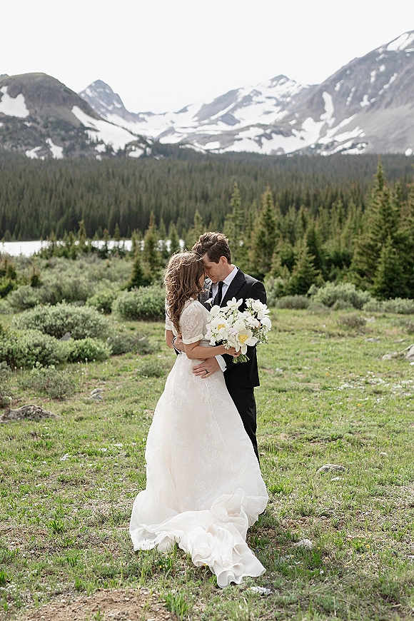 Couple portrait in a mountain wedding portrait, bride in lace gown holding a white bouquet as groom kisses her forehead by a lake meadow