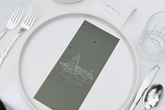 Wedding place card atop a white dinner plate with napkin, silver flatware, water glass, and menu card on a crisp white tablecloth