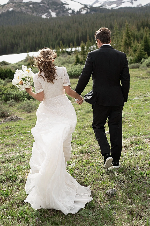 Couple portrait of bride and groom walking hand in hand, bouquet and dress train flowing by a lakeside alpine meadow with mountains