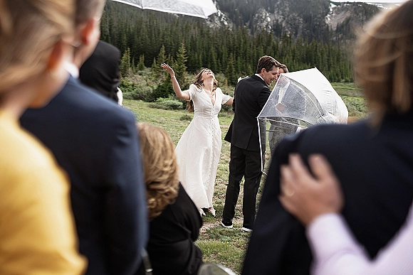 Ceremony moment at an outdoor wedding ceremony as bride in a simple dress raises arms beside groom, guests with clear umbrellas in mountain meadow