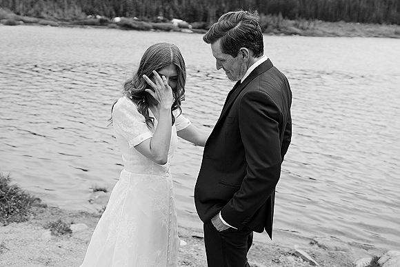 Wedding couple portrait in a black and white wedding photo, bride wiping tears as groom holds her by a mountain lake shoreline with trees