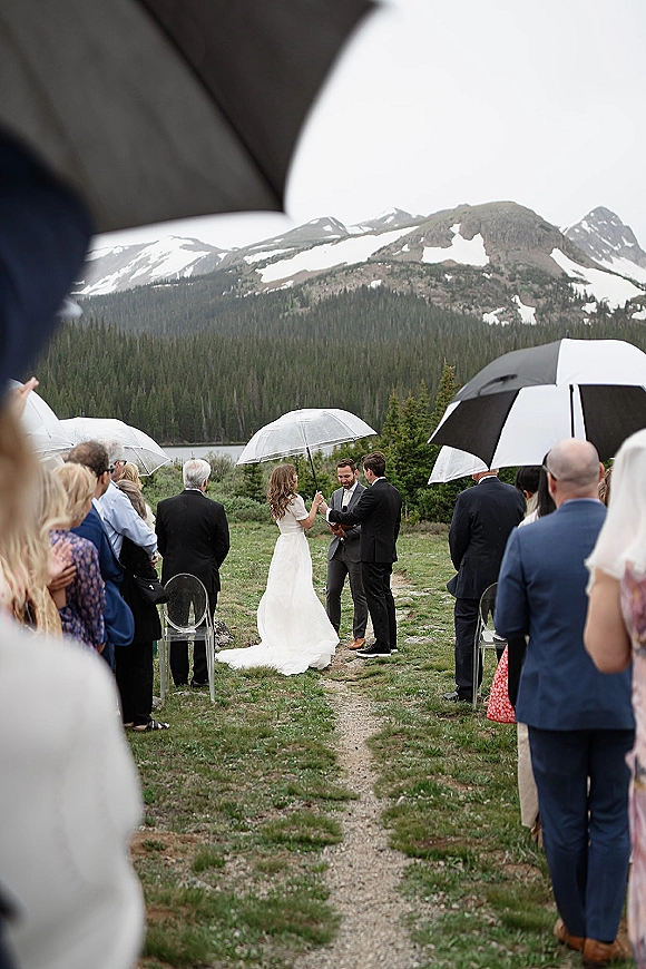 Outdoor wedding ceremony with clear umbrellas as bride and groom exchange vows in a grassy meadow by lake, mountains, and pine forest