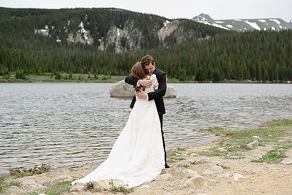 Wedding couple portrait of bride and groom embrace, her long-sleeve dress train on rocky lakeshore with pine forest and snowy mountains under overcast sky