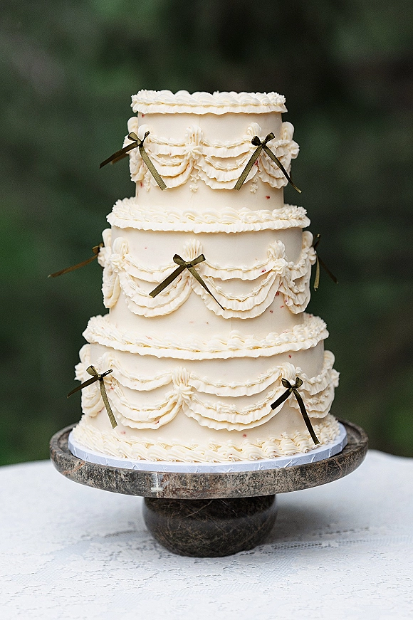 Wedding cake with white buttercream frosting, piped ruffles and ribbon bows on a cake stand, set on lace tablecloth with foliage bokeh