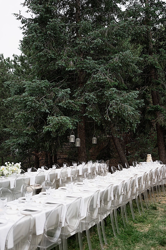 Reception tablescape for an outdoor wedding reception with long white banquet tables, clear acrylic chairs, floral centerpieces, and hanging lanterns under evergreens