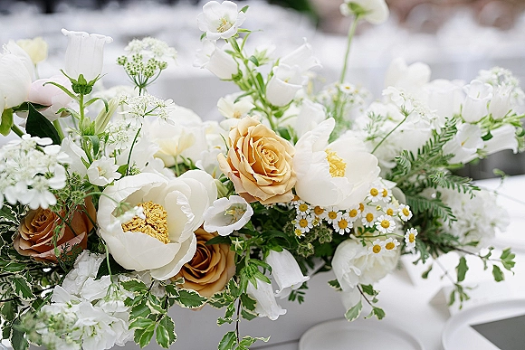Wedding centerpiece with white and peach blooms, roses, peonies, chamomile daisies, and greenery on linen-covered reception table