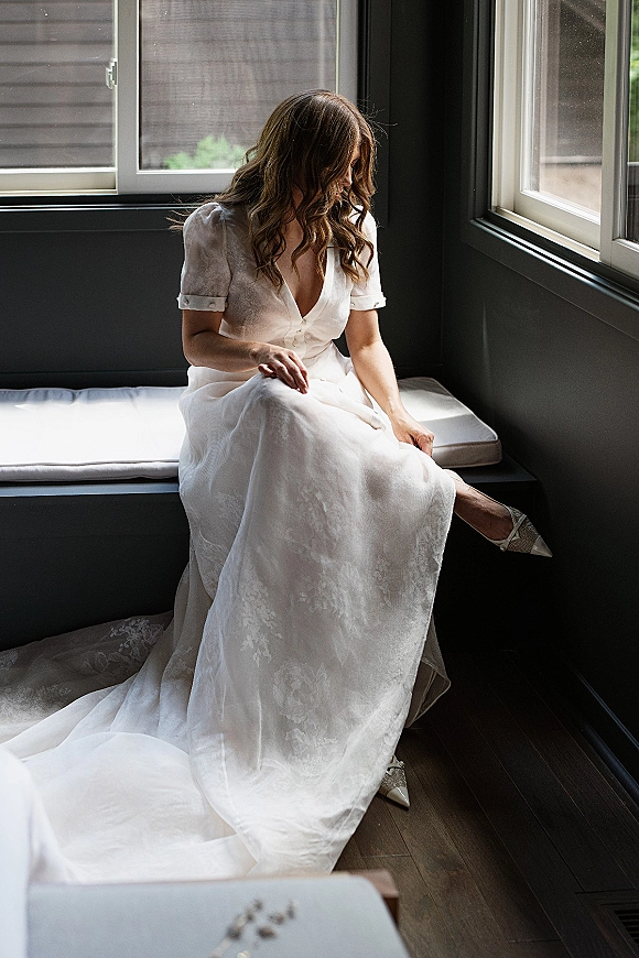 Bridal portrait of a bride sitting by window in a puff sleeve wedding dress, looking down while putting on pointed-toe heels by a window seat