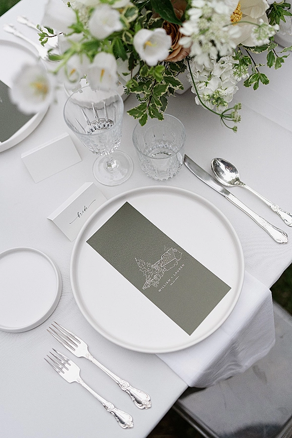 Wedding tablescape with white wedding place setting, silver flatware, crystal stemware, menu and place card on white linen beside greenery centerpiece