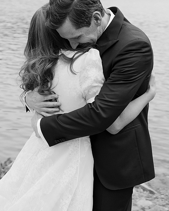 Wedding couple hug as bride and groom embrace in lace-sleeved dress and suit jacket by a calm lake shoreline backdrop