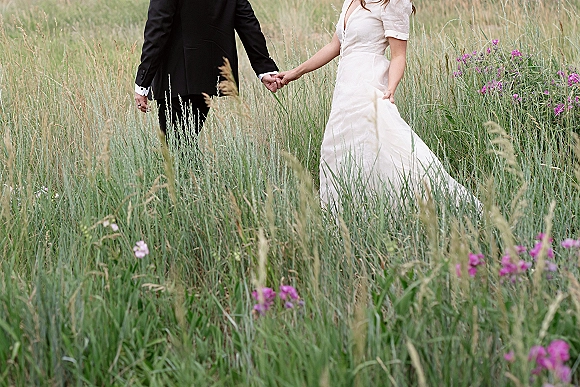 Couple portrait of bride and groom holding hands, walking through tall grass and wildflowers, her wedding dress train trailing behind