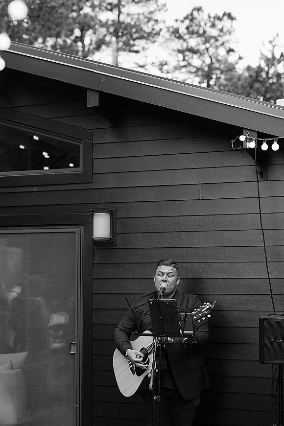Wedding musician playing acoustic guitar and singing into a microphone beside a music stand under string lights by dark wood siding