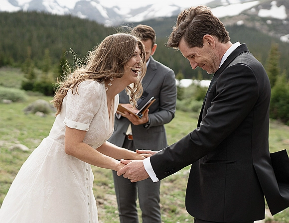 Wedding vows as bride in lace sleeves and drop earrings holds groom’s hand while officiant reads, with snowcapped mountains behind