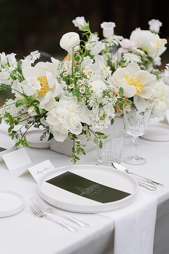 Reception tablescape with a white wedding tablescape featuring peonies and ranunculus in a compote vase, silver flatware, and menu cards in a garden setting