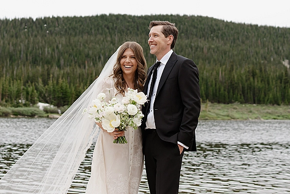 Couple portrait of a laughing bride and groom by a lakeside, bride in long veil holding a white bouquet with forested mountains behind
