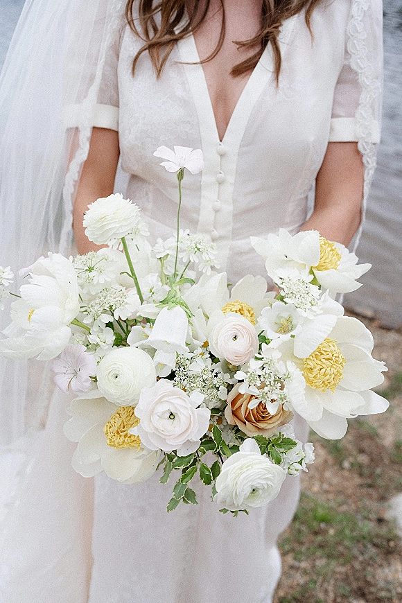 Bridal bouquet with white and yellow wedding bouquet blooms of ranunculus, anemones, roses and greenery held against a veil and button-front dress