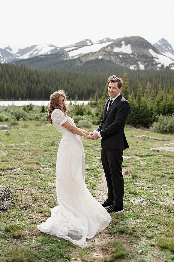 Couple portrait of bride and groom holding hands, her flowy gown and his suit, with a lakeside meadow and snowy peaks behind