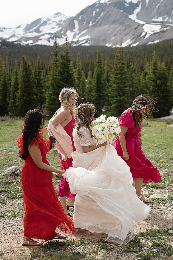 Bridesmaids with bride walking a dirt path, holding her dress train as she carries a white flower bouquet in a windy mountain meadow backdrop