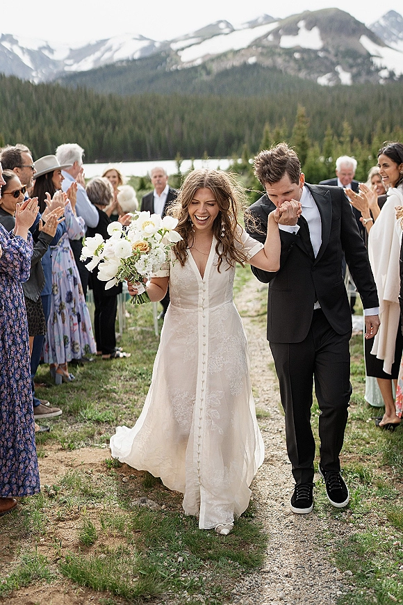 Wedding recessional as bride and groom walk down the aisle, groom kissing her hand, bouquet in hand, guests cheering by lake and pines