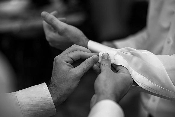 Groom getting ready as hands fasten cufflinks on a white dress shirt cuff, focusing on buttons and details in a blurred indoor room
