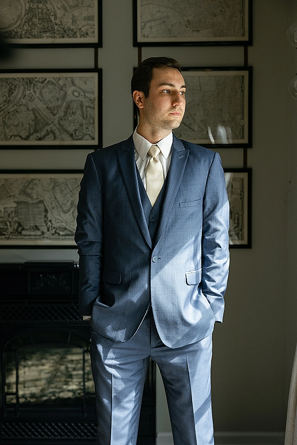 Groom portrait in a blue wedding suit, hands in pockets by window light, with framed wall art and dark bench behind him