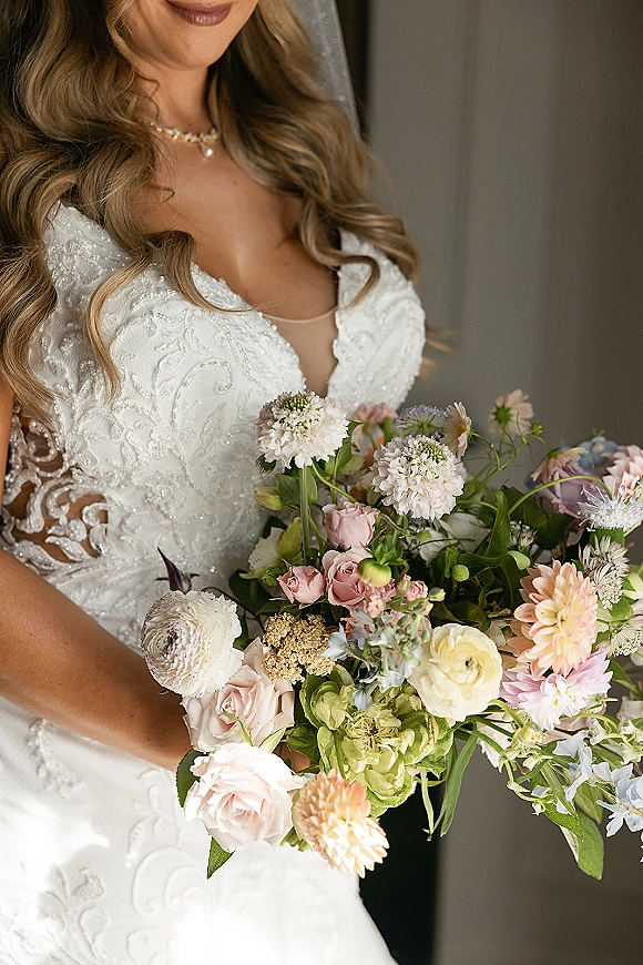 Bridal portrait of a bride holding bouquet of blush roses, dahlias, and ranunculus, wearing a lace dress and pearl necklace in window light