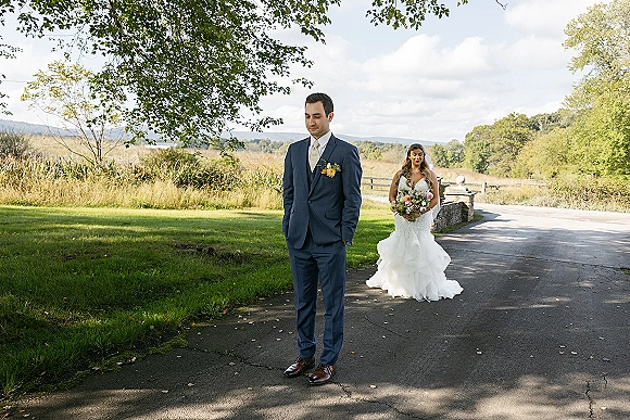 Wedding first look as bride in a veil approaches groom from behind on a country road, holding a colorful bouquet by a rustic fence