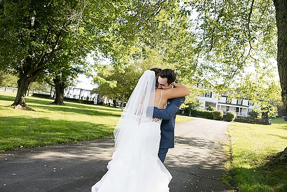 Wedding couple portrait of bride and groom hug on a sunlit tree-lined path, her lace gown and veil visible beside a farmhouse backdrop