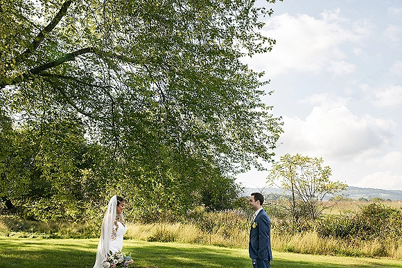First look moment outdoors as bride in lace gown with veil and bouquet faces groom in blue suit under tree canopy in a wild meadow