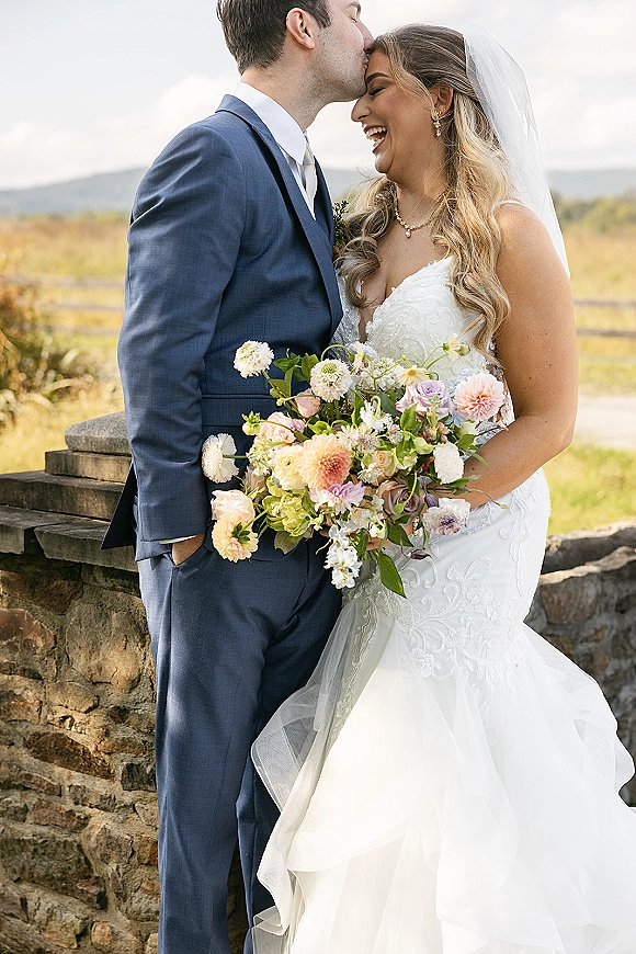 Couple portrait of groom in a navy suit kissing the bride’s forehead as she holds a bouquet, with veil and stone wall countryside backdrop
