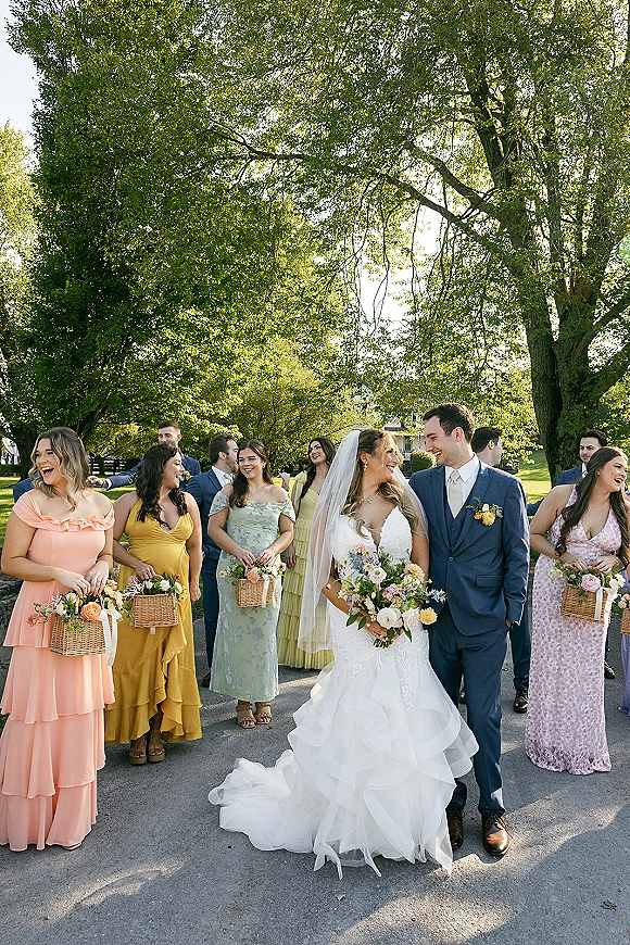 Wedding party portrait of bride and groom with wedding party walking along a sunlit tree-lined path, bridesmaids in pastel dresses with baskets