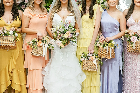 Bridesmaid group photo with bride and bridesmaids in pastel dresses holding wicker basket bouquets before a floral greenery backdrop