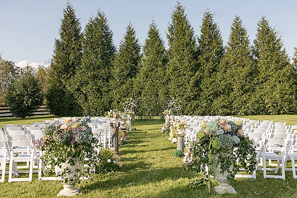 Outdoor ceremony setup with wedding ceremony seating, white folding chairs lining a flower-filled aisle on a lawn framed by evergreens and blue sky