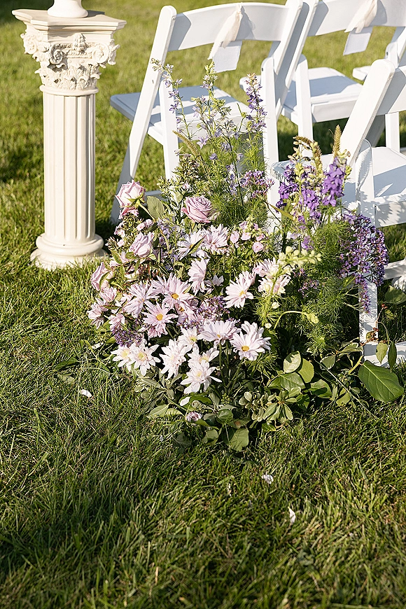 Ceremony aisle flowers in a wildflower cluster of roses, daisies, and purple blooms beside white folding chairs on a grass lawn outdoors