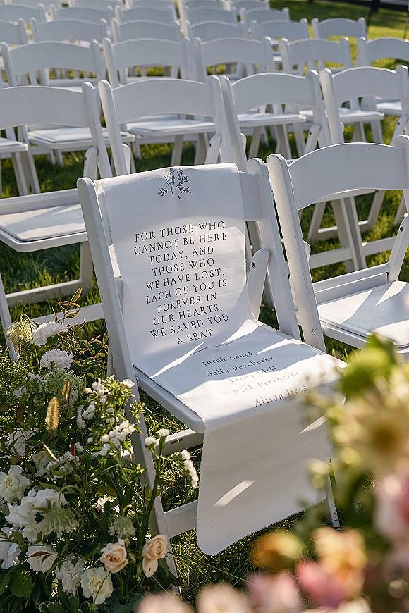 Wedding memorial seat with a reserved sign on a white folding chair, framed by blush roses and greenery along outdoor ceremony rows