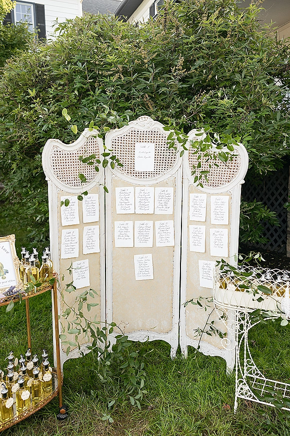 Wedding seating chart with calligraphy escort cards on a vintage folding screen, accented with greenery garland beside a gold bar cart on lawn