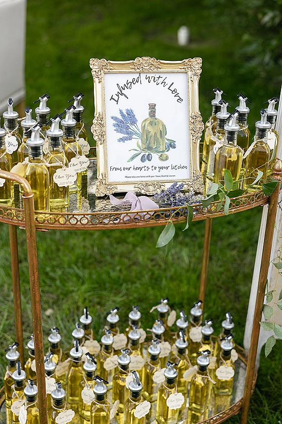 Wedding favors display with olive oil wedding favors in glass bottles, twine tags, lavender sprigs, and gold framed sign on a bar cart outdoors
