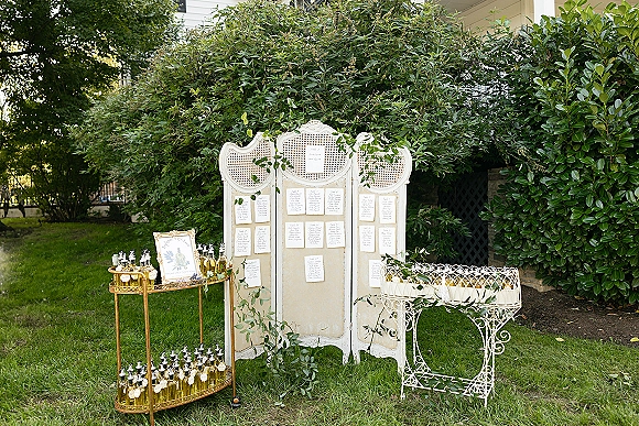 Wedding seating chart with calligraphy escort cards on a vintage folding screen, greenery garland, and gold bar cart on a garden lawn