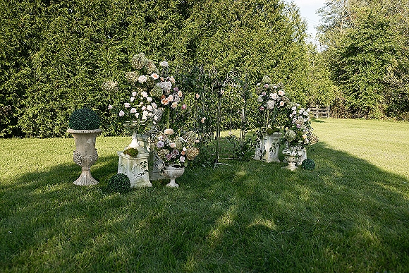 Wedding altar decor with a garden ceremony backdrop, featuring a flower-covered gate with roses and hydrangeas on a manicured lawn