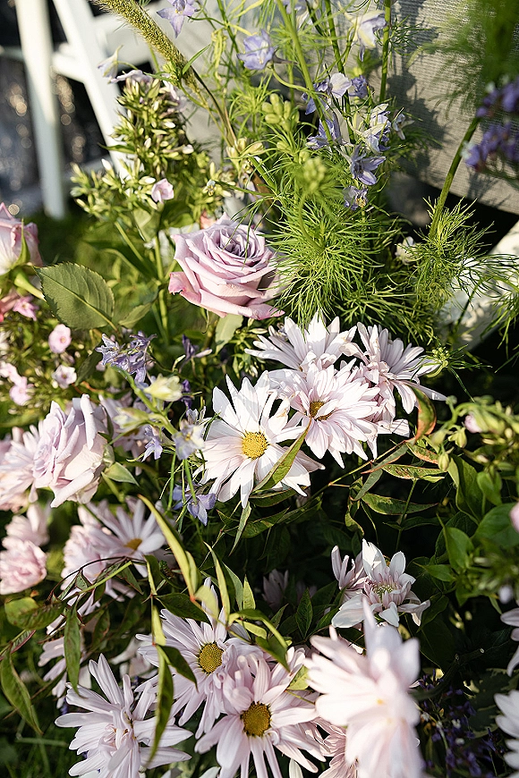 Wedding floral arrangement with lavender roses and white daisies nestled in greenery beside white folding chairs on a grass lawn