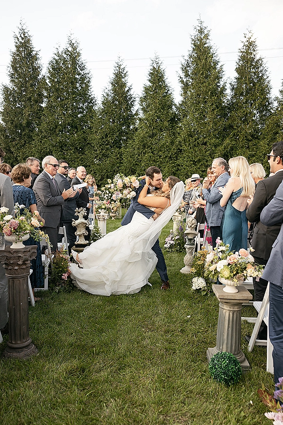 Wedding kiss moment as groom dips bride in flowing veil and gown, pastel aisle florals and cheering guests on a green lawn with evergreens
