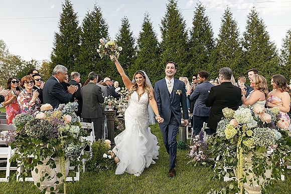 Wedding recessional with bride and groom walking aisle, bride raising bouquet as guests cheer beside hydrangea-lined chairs on lawn