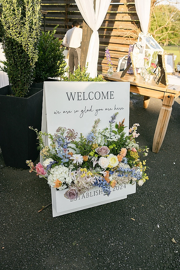 Wedding welcome sign with flowers in elegant calligraphy, set on a wood table with framed signs and greenery against a rustic wooden wall backdrop