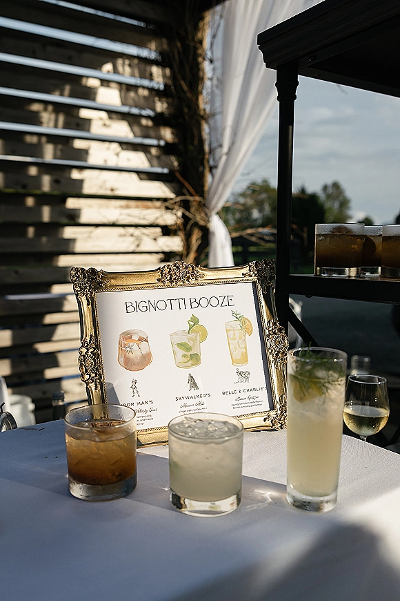 Signature drink sign in an ornate gold frame showing wedding signature cocktails, set on a bar cart with glasses against draped wood slats outdoors