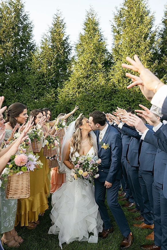 Wedding kiss portrait of bride and groom walking as the newlywed kiss aisle forms with cheering wedding party on a green lawn, veil flowing
