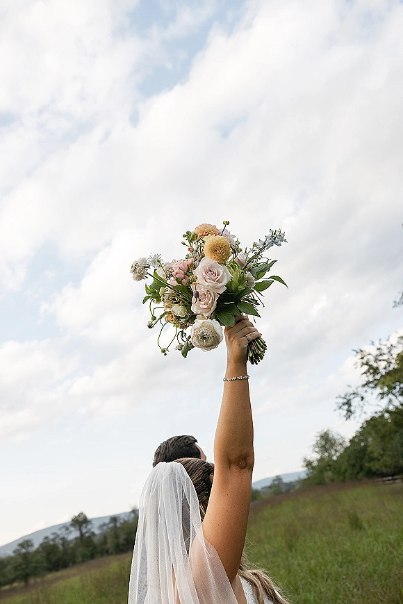 Wedding bouquet of blush roses and wildflowers raised in the air, with greenery accents against open sky and meadow hills