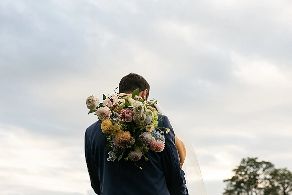 Wedding couple portrait of bride and groom hug, with bouquet over shoulder and veil, groom in navy suit under cloudy sky and trees