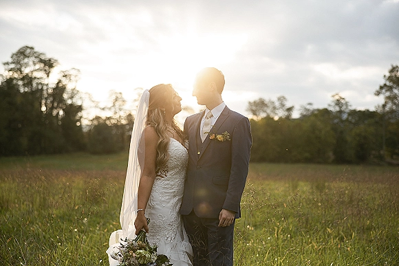 Couple portrait of bride and groom at sunset, her veil and bouquet glowing beside his suit and boutonniere in a meadow with trees