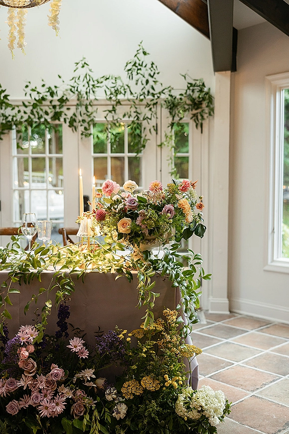 Reception sweetheart table with sweetheart table florals, greenery garland, taper candles and ribbed glass holders beneath overhead greenery.