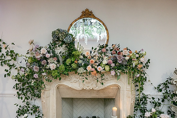 Wedding fireplace decor with a rose and hydrangea floral garland draped on a carved mantel beneath a round mirror, plus a pillar candle