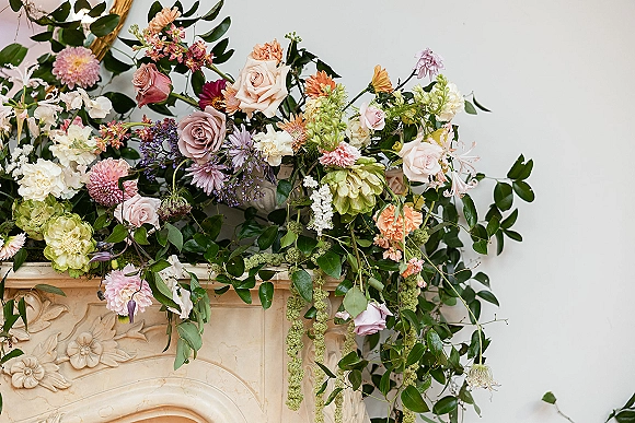 Wedding floral installation draped over a stone mantel with pastel roses, dahlias, carnations, and trailing vines beneath a mirror frame